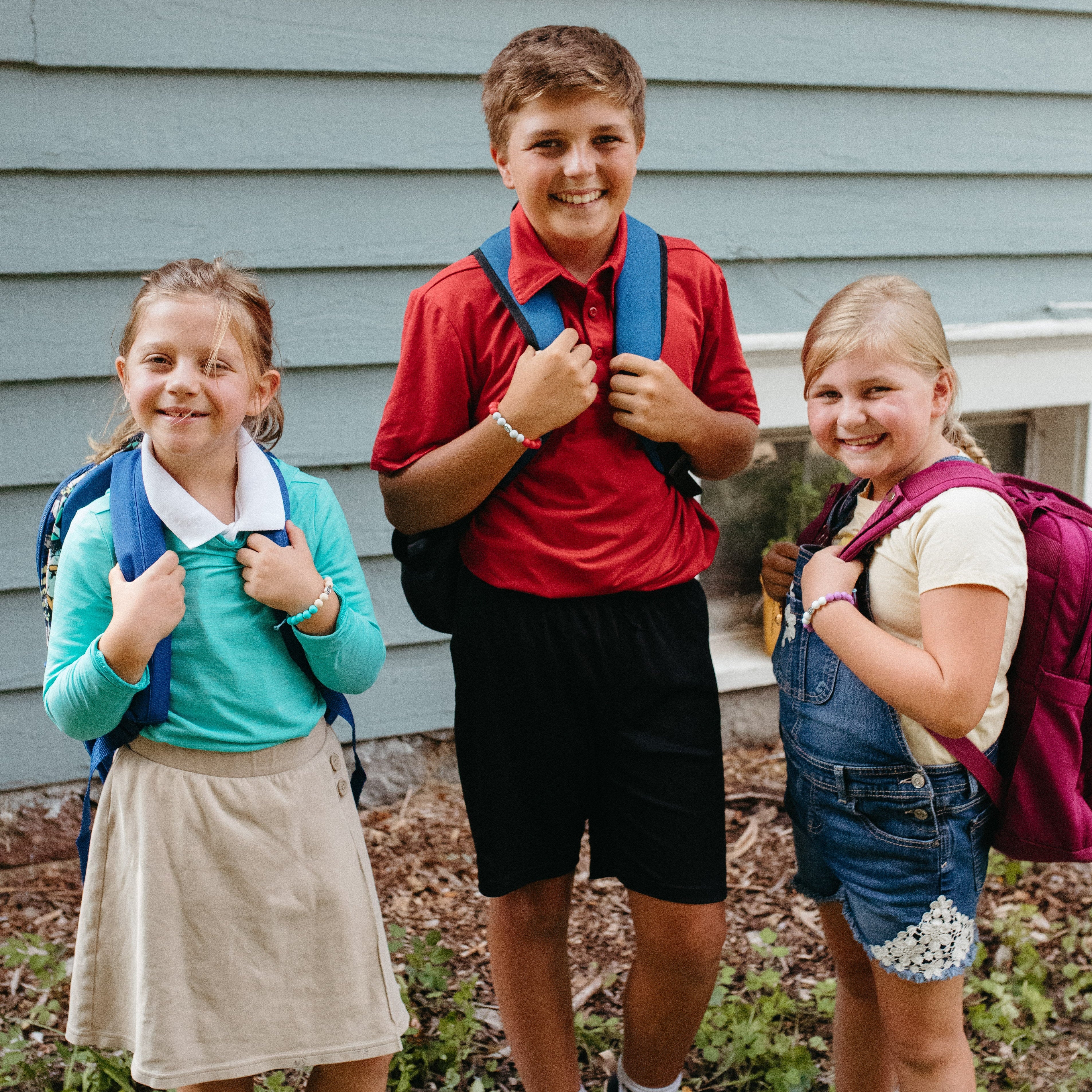 Three children standing outdoors in front of a light blue house, each wearing a backpack. The child on the left wears a turquoise top and beige skirt, the center child wears a red polo shirt and black shorts, and the child on the right wears denim overalls and a light top.