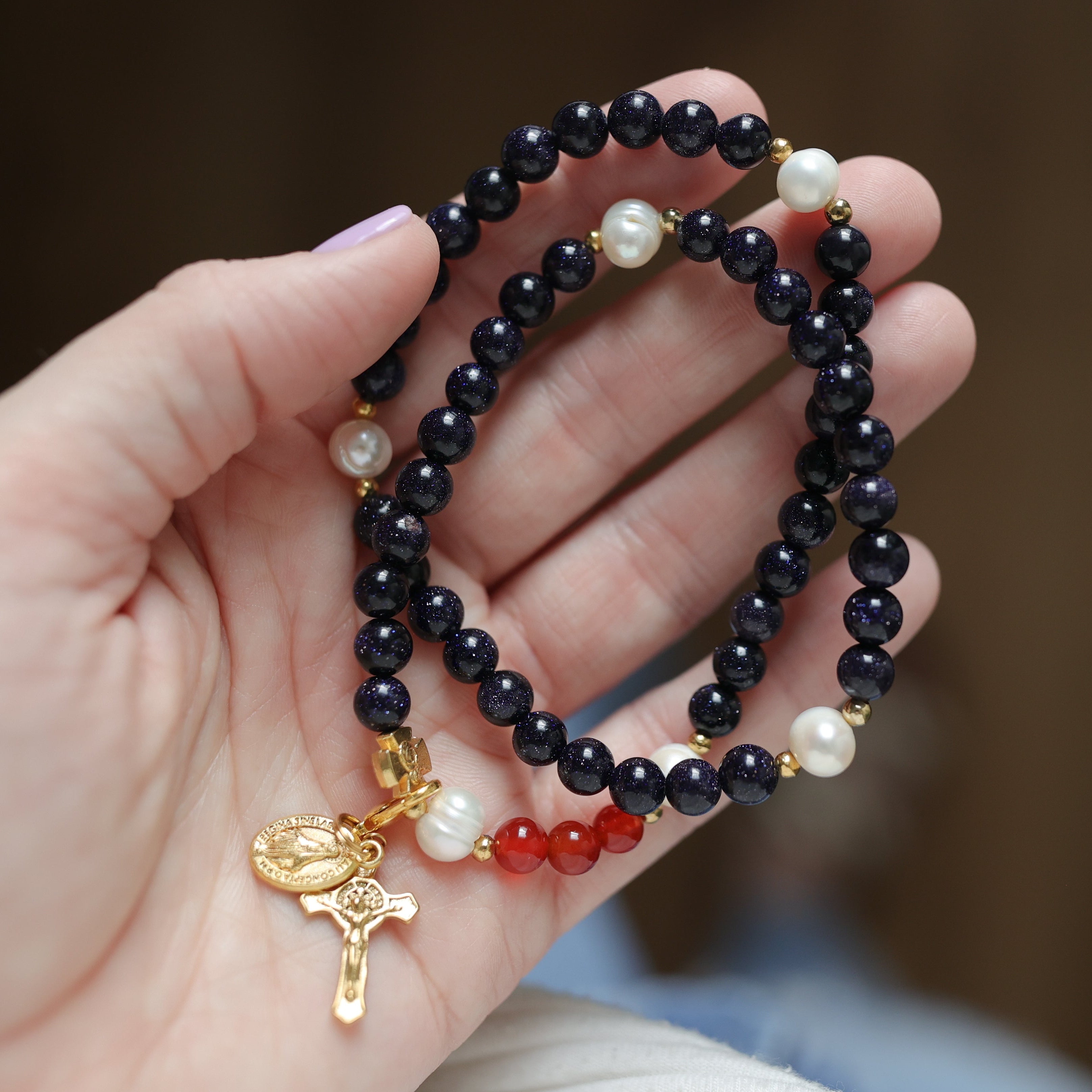 Hand holding a dark blue beaded bracelet with gold and red accents against a blurred background