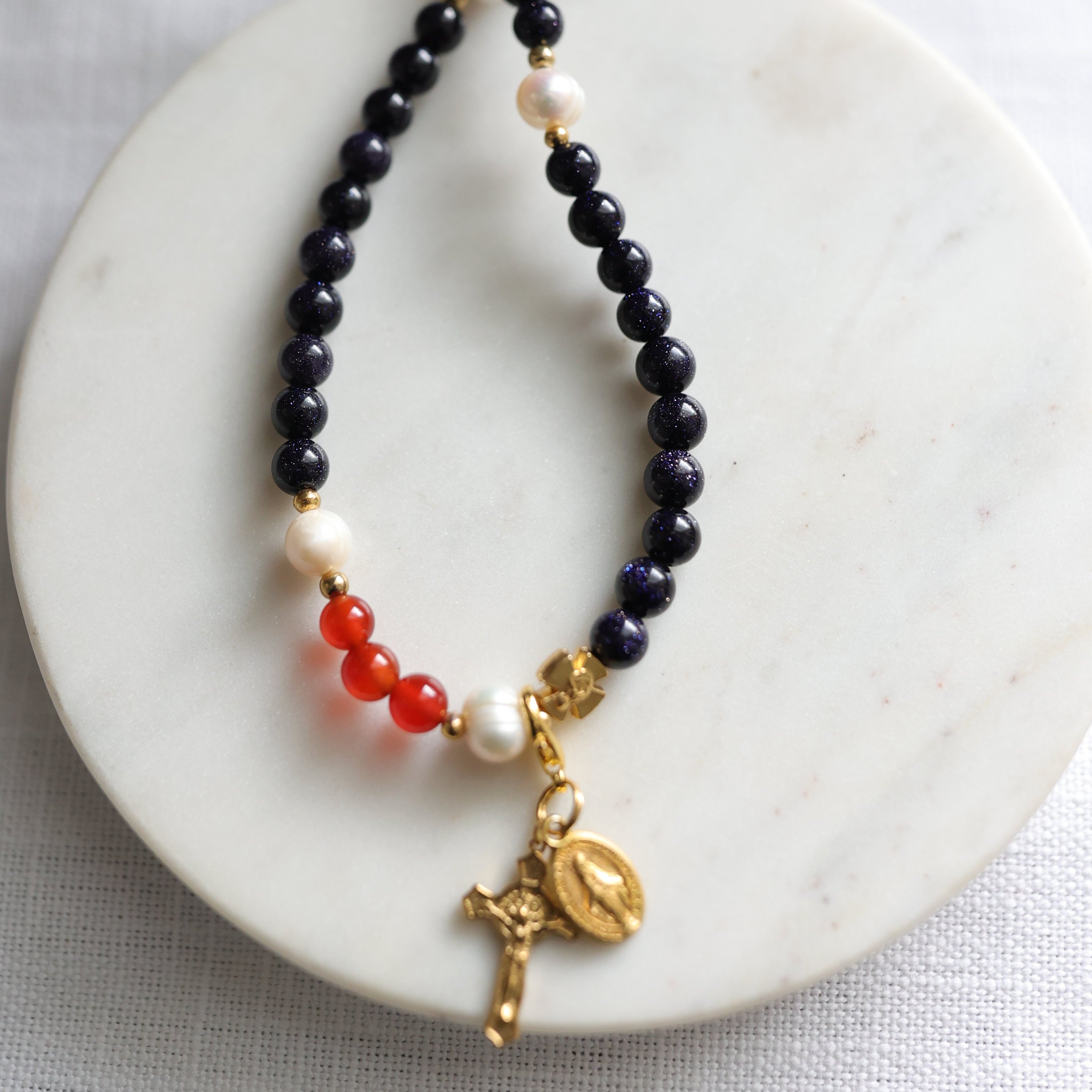 Dark blue gemstone and peark beaded rosary with gold and red charms on a white marble surface