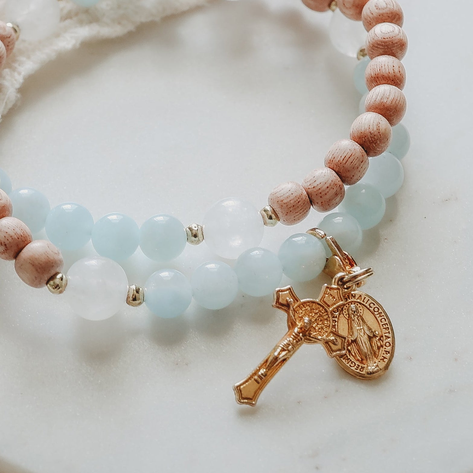 A beaded stretch bracelet with light blue and peach beads, a small gold cross charm, and gold spacer beads, displayed on a round white marble coaster. The scene is styled with a cream lace cloth, green leaves, and small white flowers on a wooden surface.