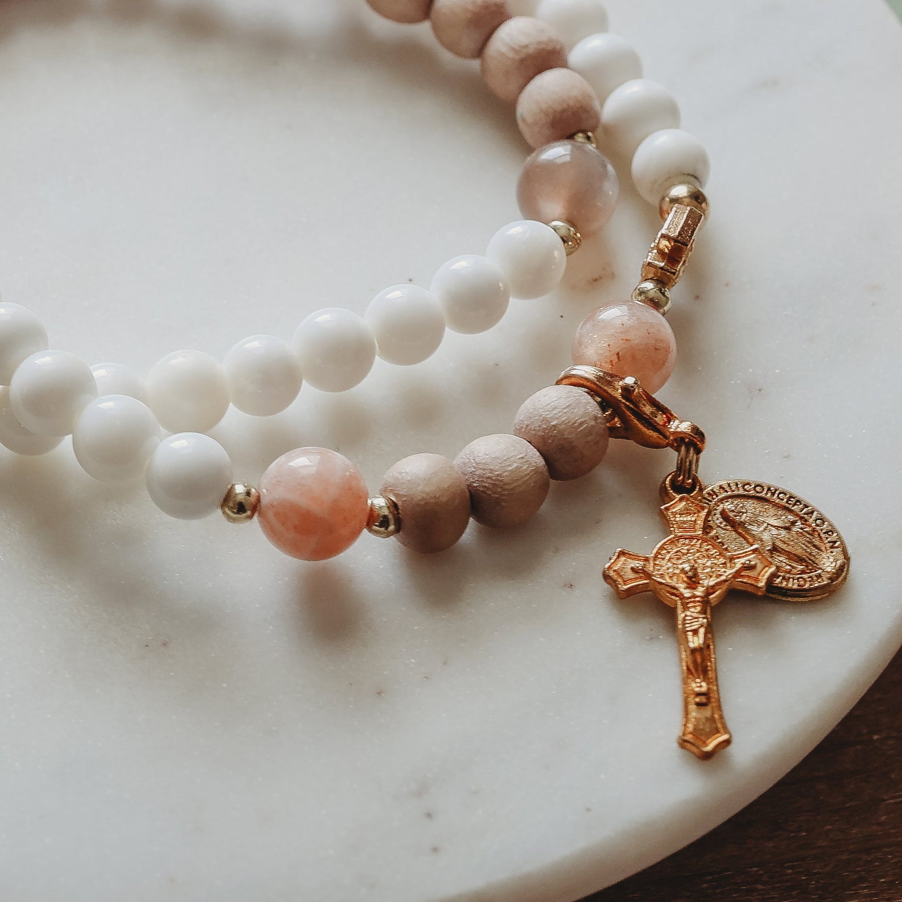 Two beaded prayer bracelets with white and light brown beads, accented with peach-colored stones and a gold-tone cross charm, displayed on a white marble dish over a lace cloth on a wooden surface.