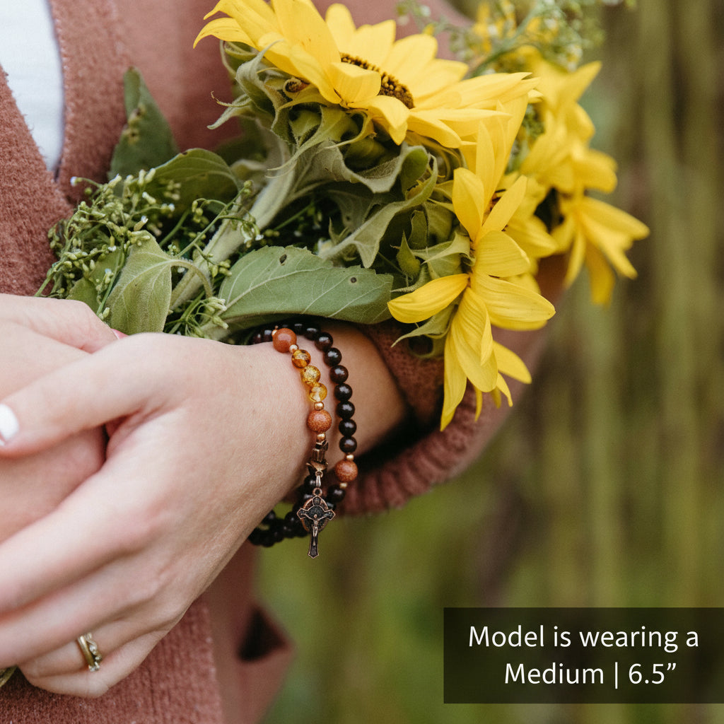 A close-up of a person's hands and torso holding a bouquet of yellow sunflowers outdoors. The person is wearing a rust-brown knit sweater and several beaded bracelets on one wrist, including a small cross charm.