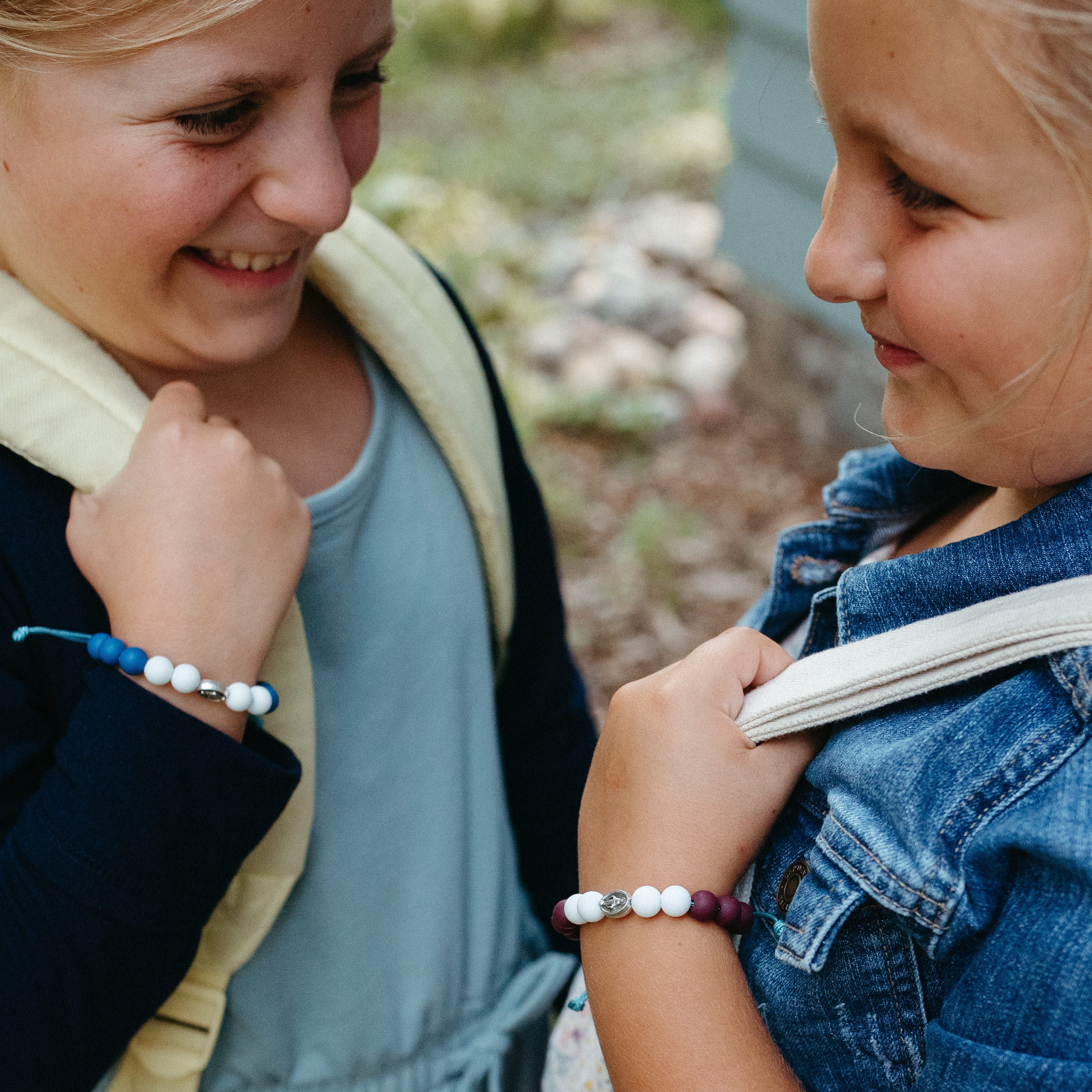 Two young girls smiling at each other outdoors, each wearing a beaded bracelet on their wrist. One bracelet is blue and white, and silver and the other is purple, white, and silver.