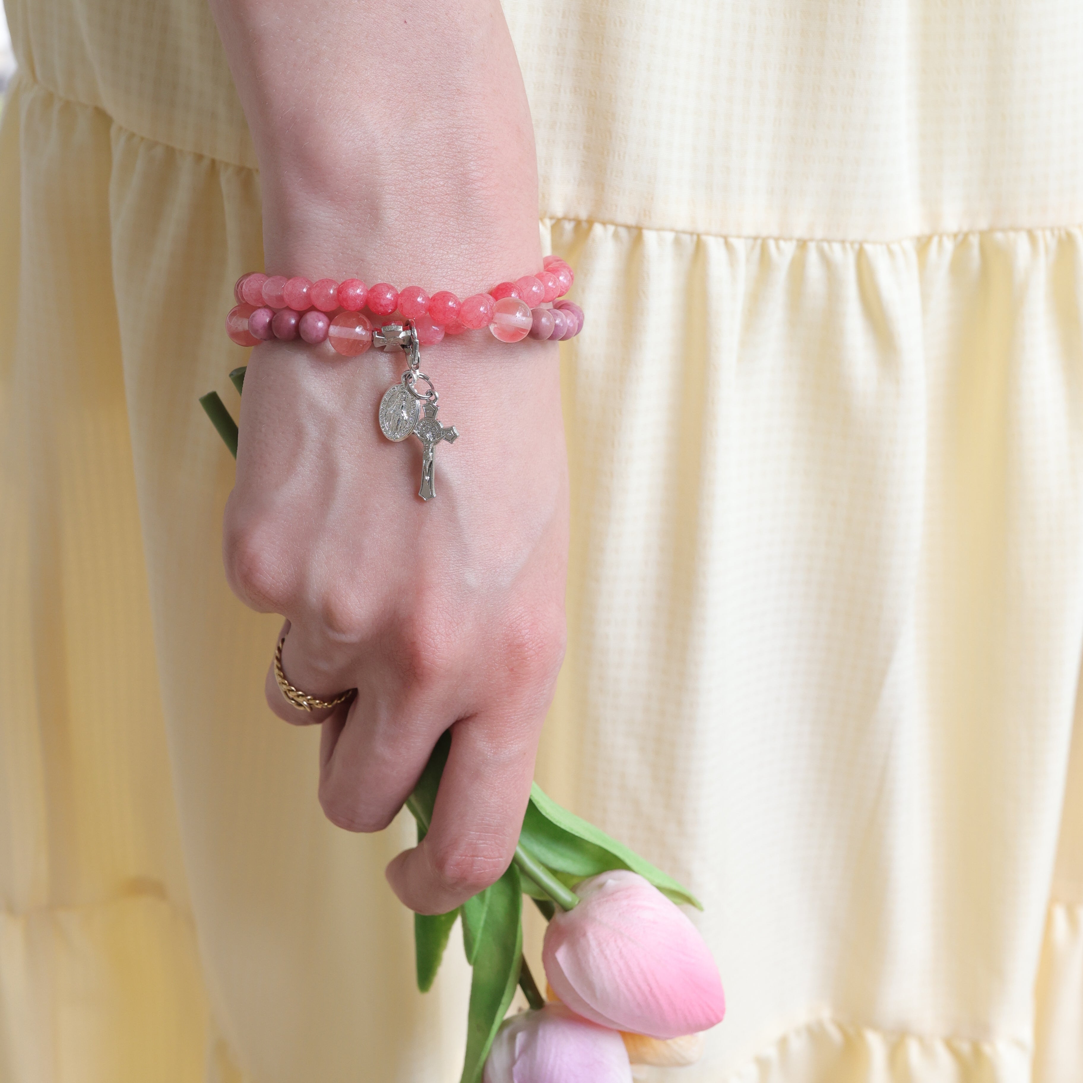 Person wearing a pink beaded bracelet with a cross charm, holding pink tulips against a yellow background.