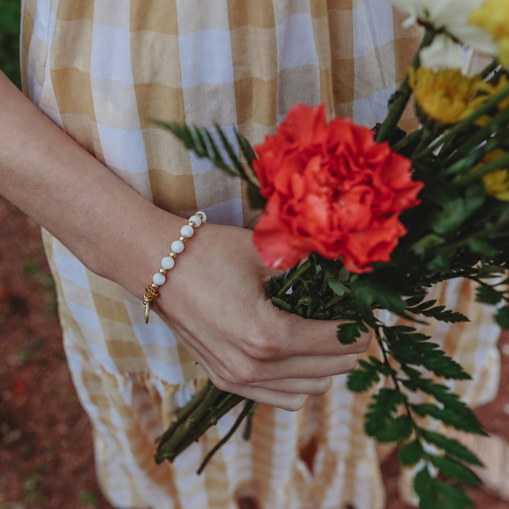 A close-up lifestyle shot of a person holding a vibrant bouquet of flowers, including a prominent red carnation and yellow mums, while wearing a gold and white checkered dress and a pearl beaded bracelet.