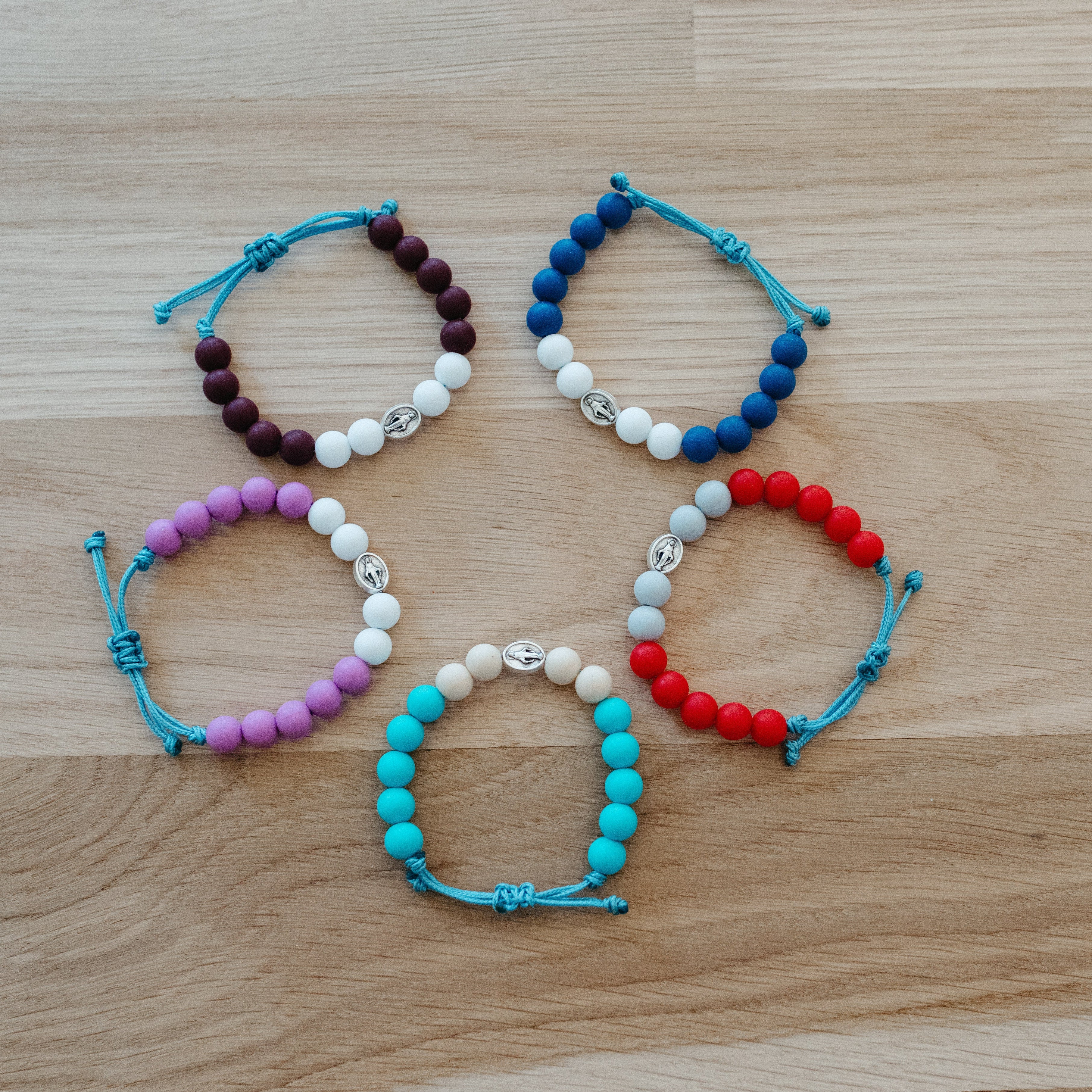 Five adjustable cord beaded bracelets arranged in a circle on a light wood grain surface. The bracelets feature different bead color combinations: dark brown, white, blue; purple, white, red; purple, white, turquoise; turquoise, white, red; and dark brown, white, blue.