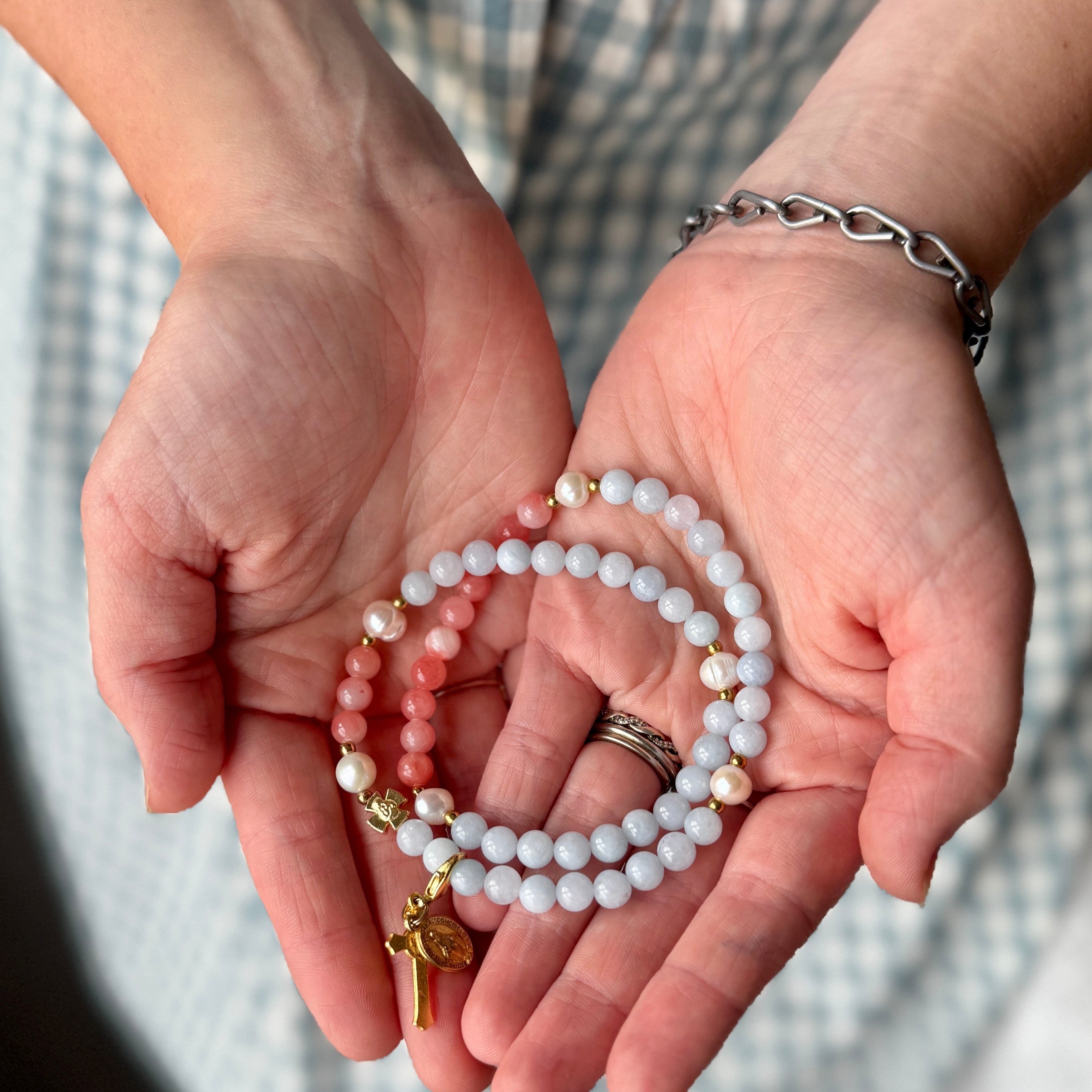 A close-up of two hands holding a set of beaded bracelets. The bracelets consist of white and pale pink round beads with small gold spacer beads and a gold cross charm.