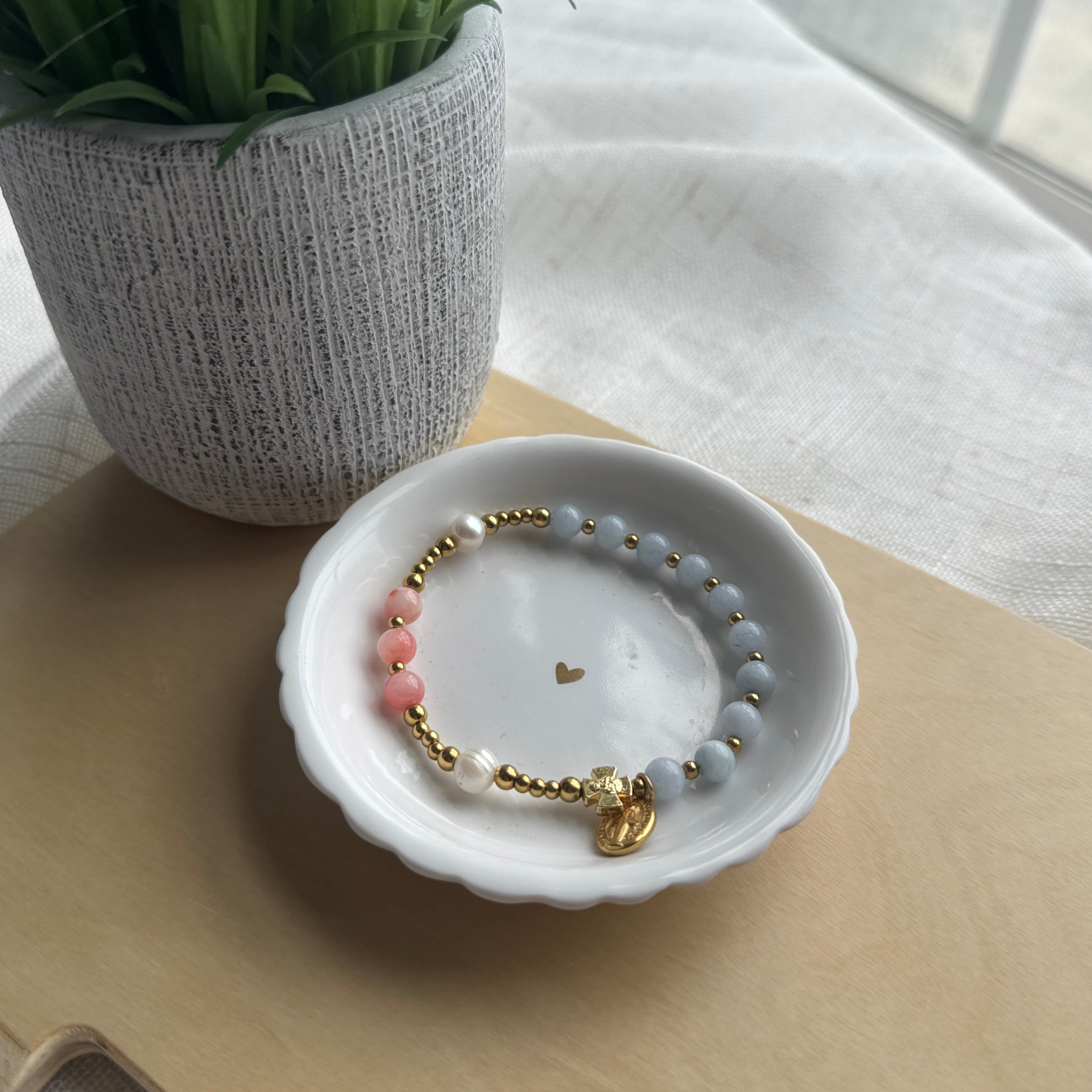 A beaded bracelet with light blue-gray beads, pink beads, white pearl-like accents, and gold-tone metal details, displayed in a small white dish on a beige surface with a potted plant in the background.