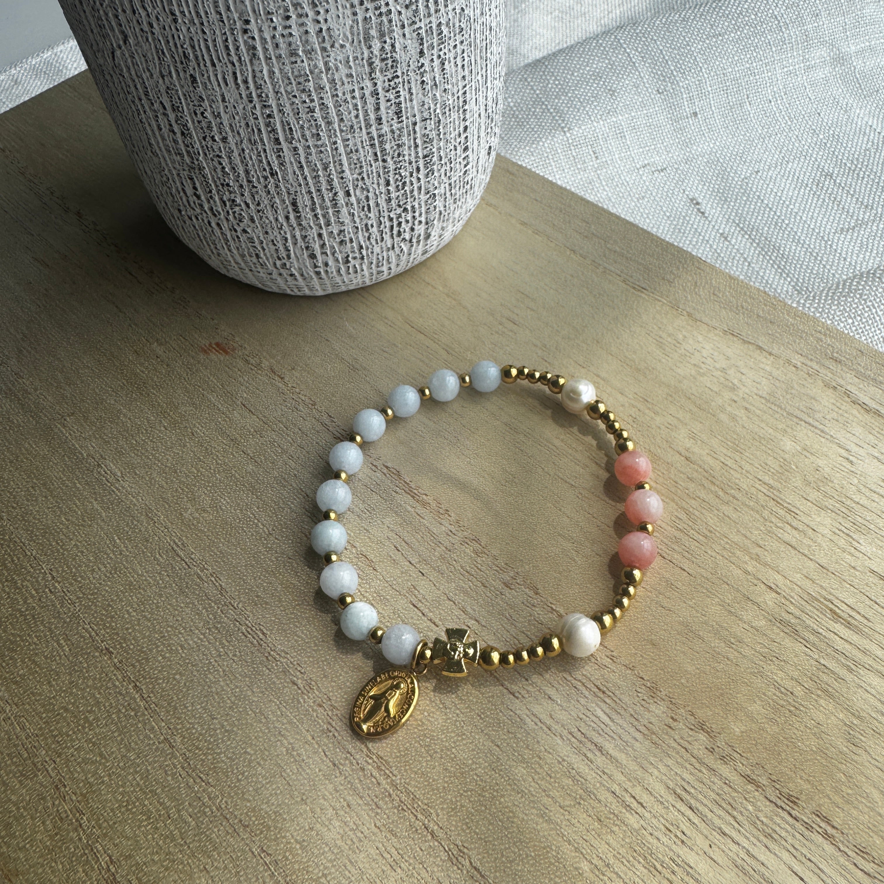 A beaded bracelet with white pearl-like beads, pink stone beads, gold spacer beads, and a small gold charm, displayed on a light wood surface.