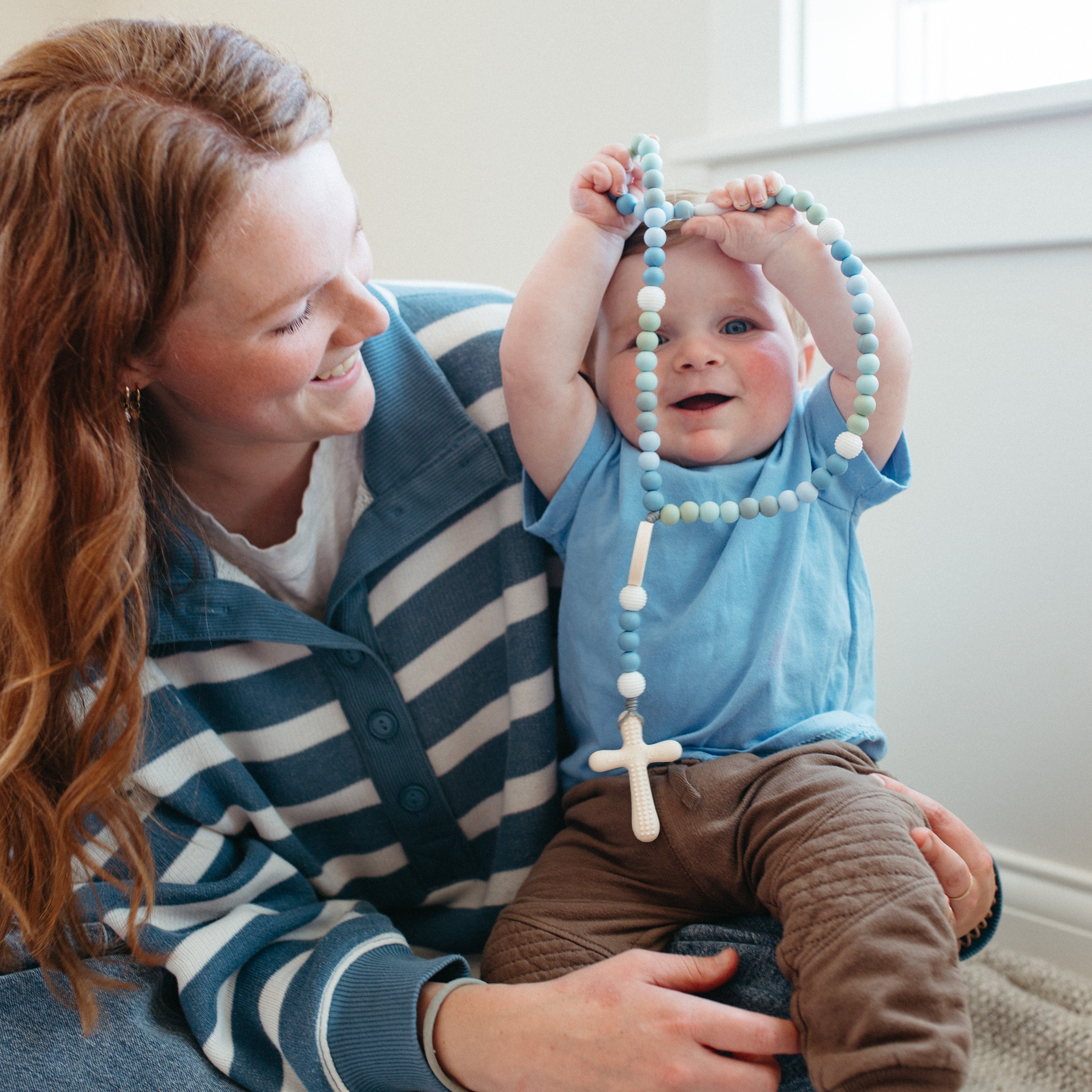 Woman holding a baby with a rosary, sitting on a carpeted floor.