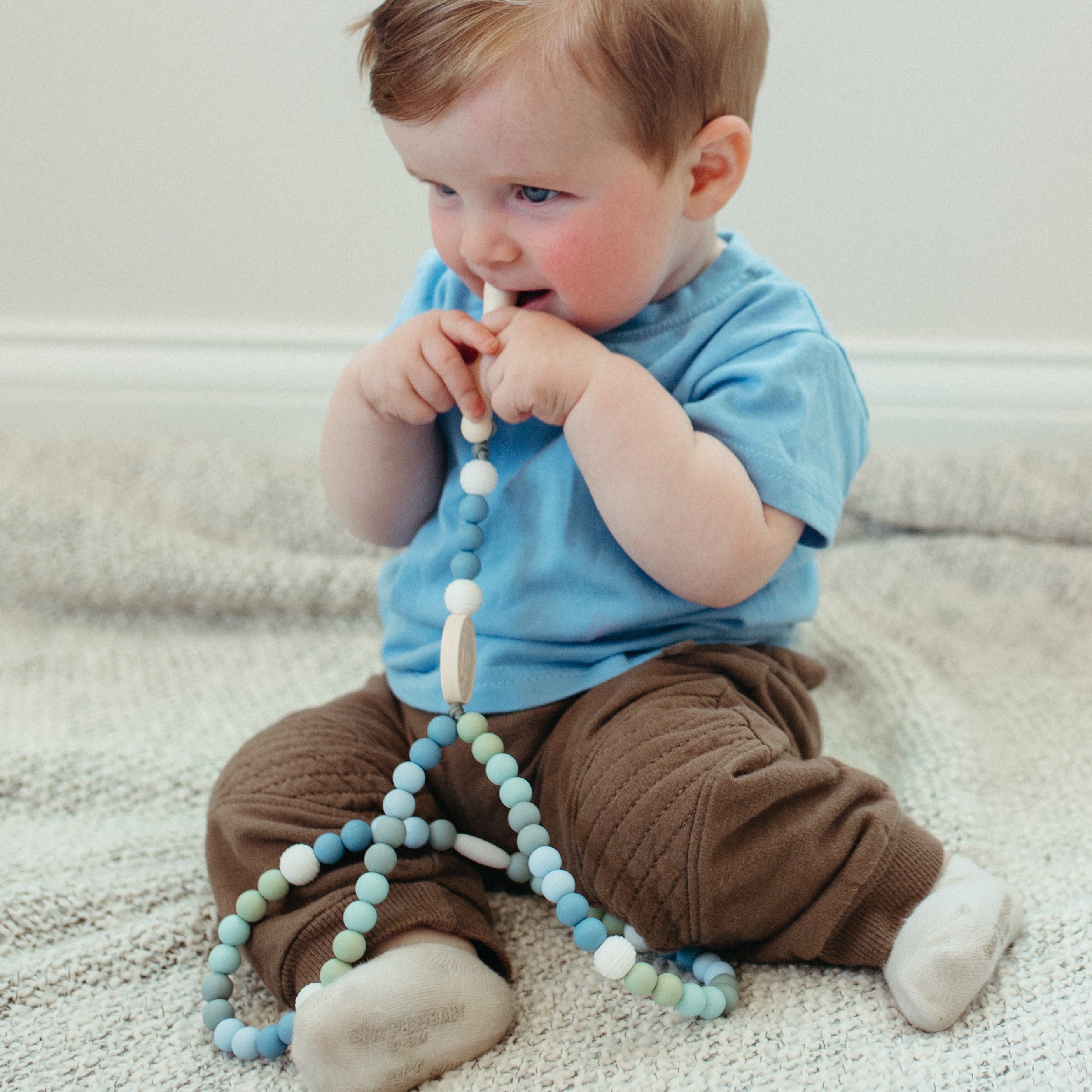 Baby sitting on a carpeted floor holding a silicone rosary.