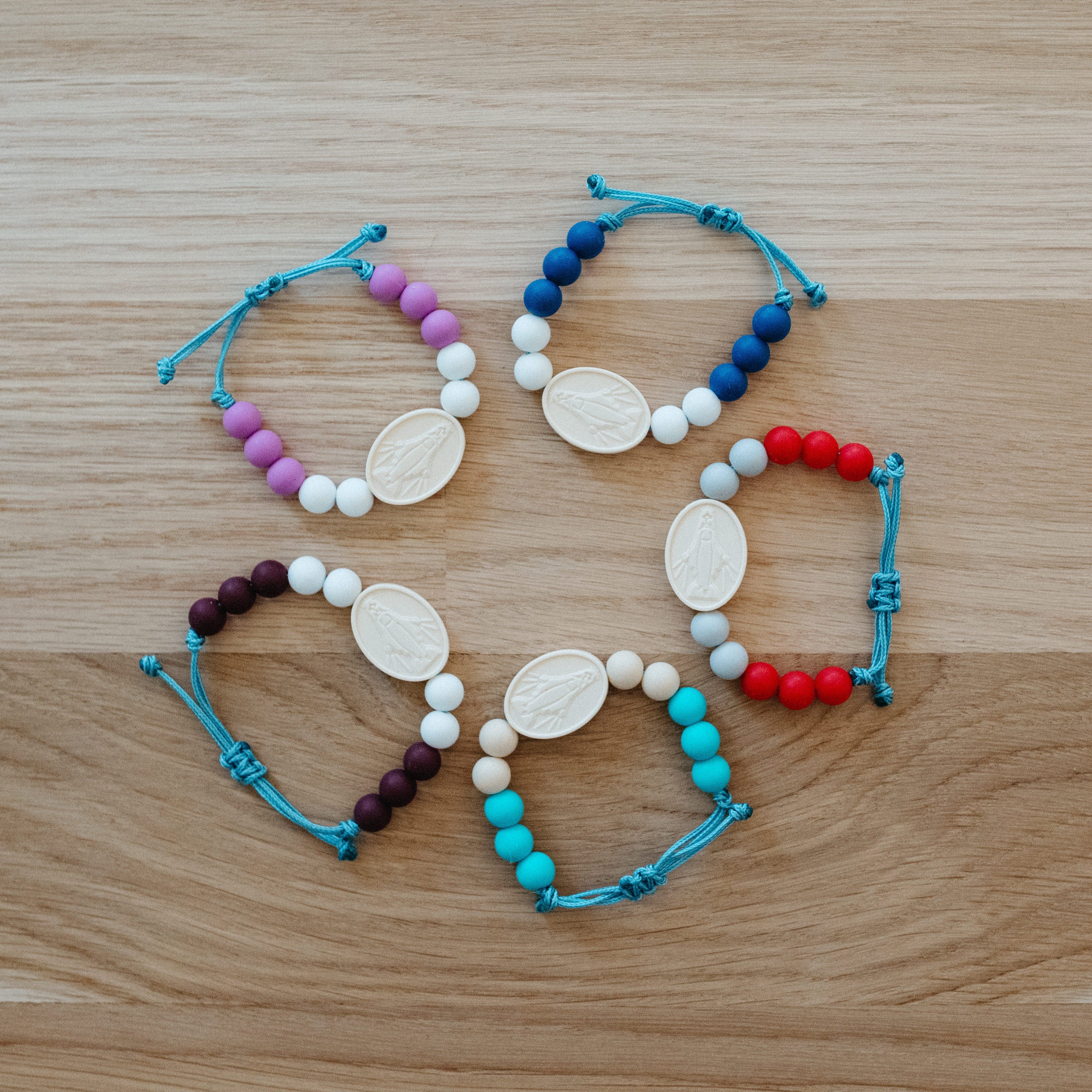 Six beaded bracelets arranged in a circle on a light wood surface. Each bracelet features white oval beads, white round beads, and a string of beads in different color combinations: blue, purple, red, dark purple, turquoise, and navy.