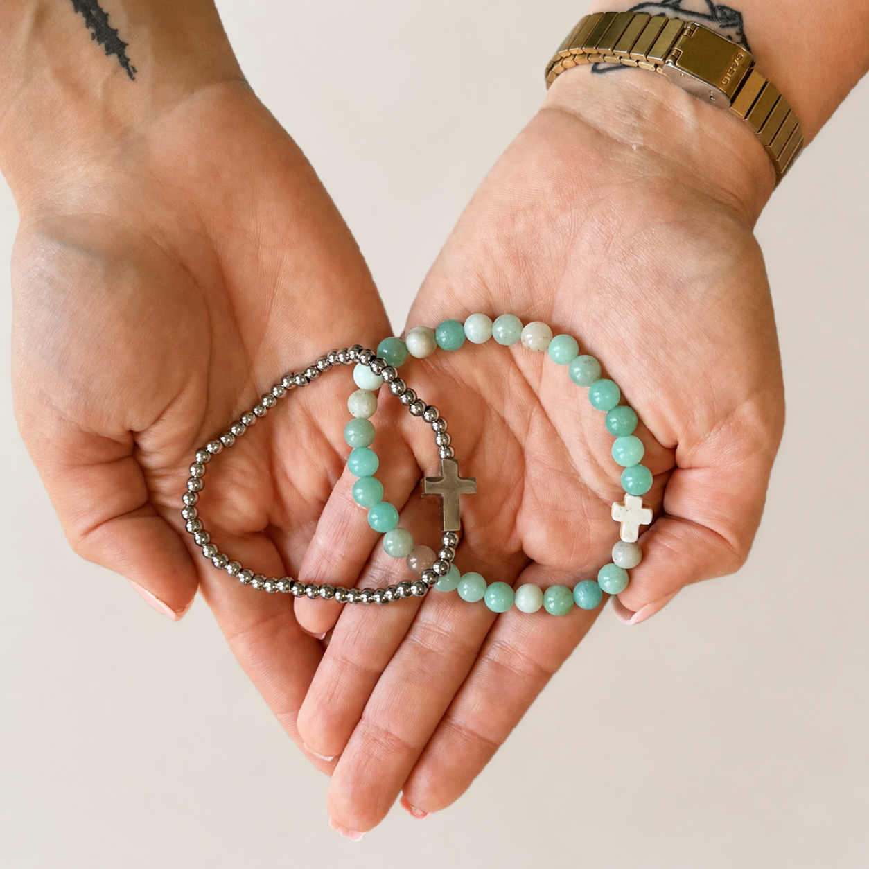 Two beaded bracelets with crosses held in a person's hands. One bracelet has light mint green and white beads with a silver cross, and the other has silver-toned beads with a white cross.