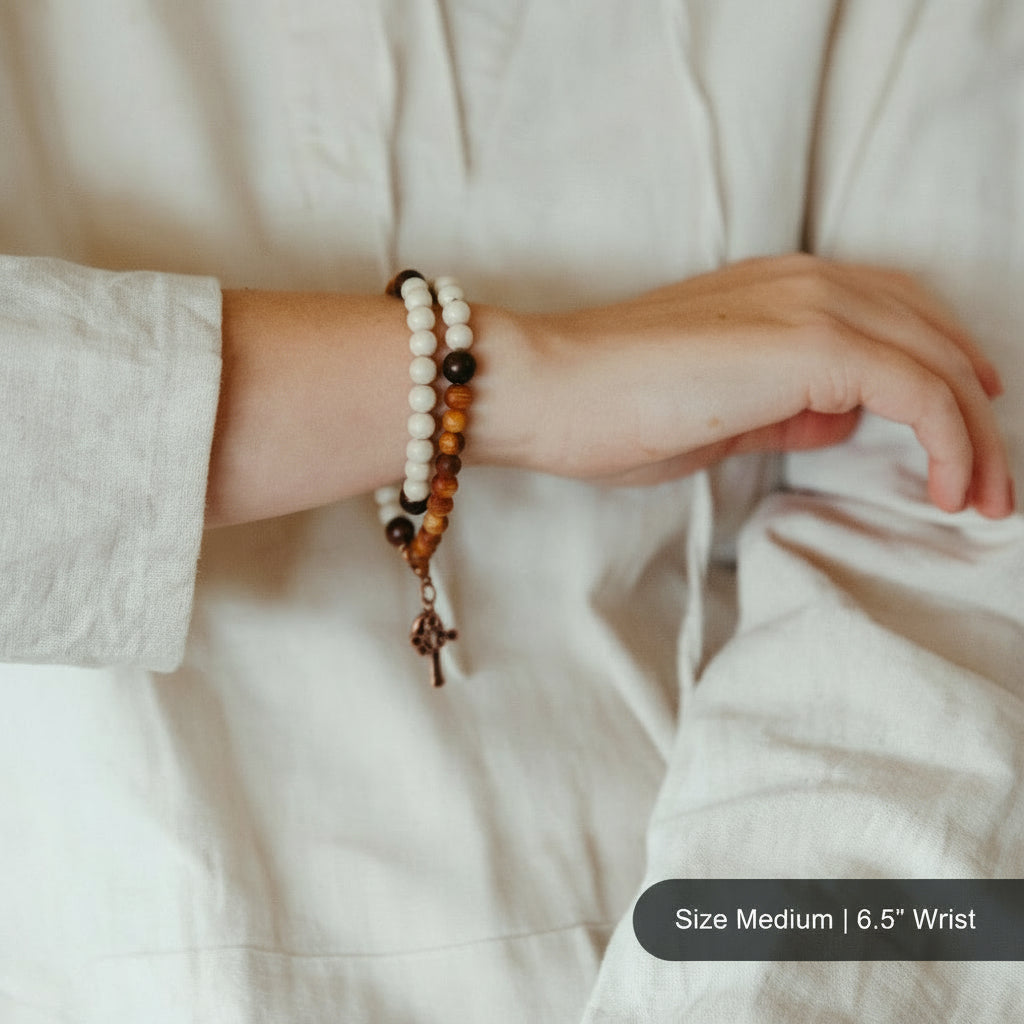 A close-up of a person wearing a double-strand beaded bracelet with white and brown beads and a small wooden cross, styled with a light beige linen shirt.