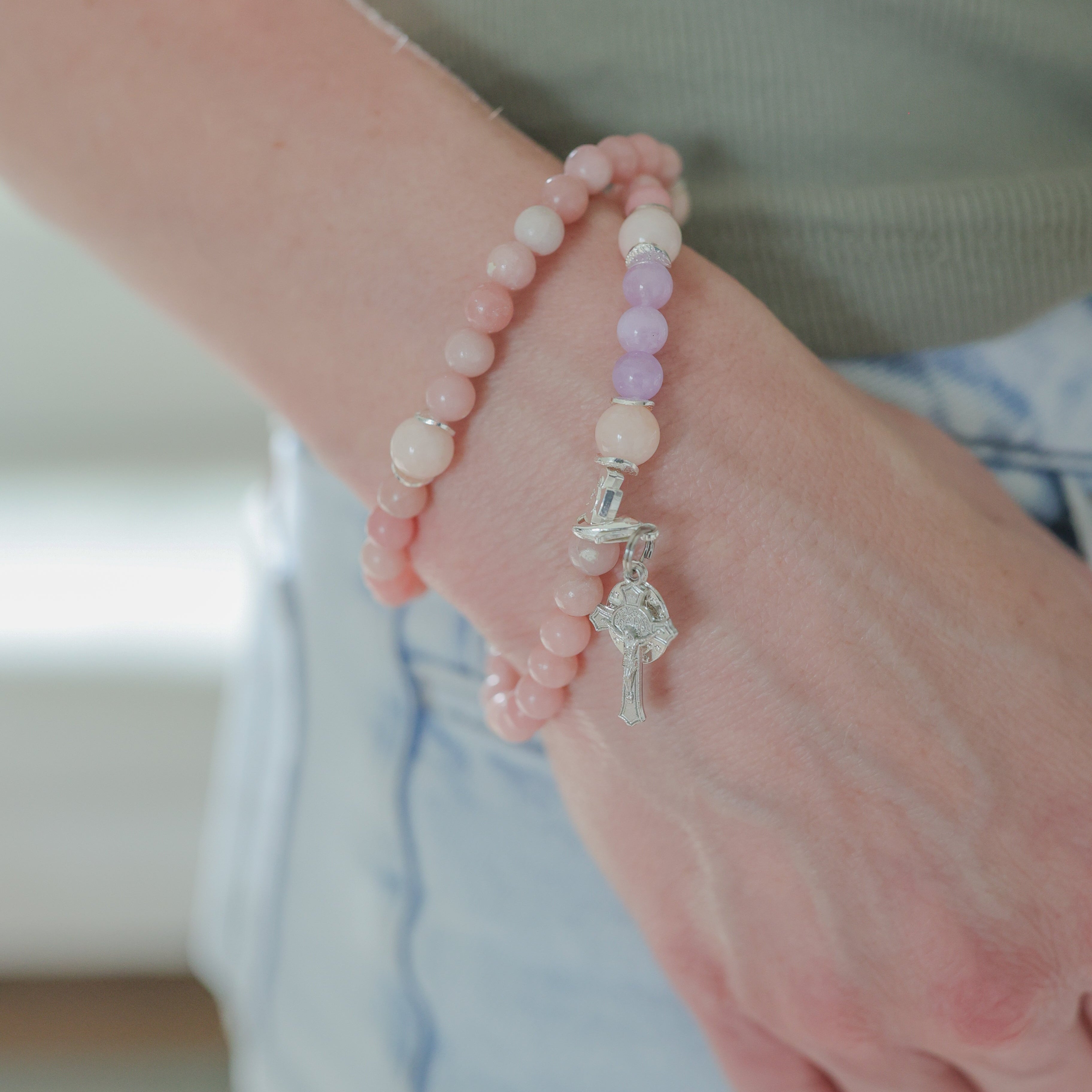 A close-up of a person's wrist wearing two beaded bracelets. One bracelet is made of light pink beads with a silver-toned fairy charm, and the other is made of alternating pastel pink and lavender beads with a silver-toned fairy charm.